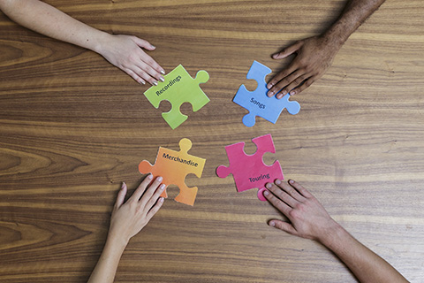 Four individual hands are holding four different colored puzzle pieces with the words recordings, songs, merchandise, and touring printed on them